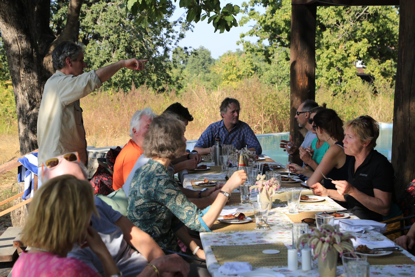 Guests at a long lunch table beneath the trees by the lodge pool in Pench, as the naturalist points towards the forest
