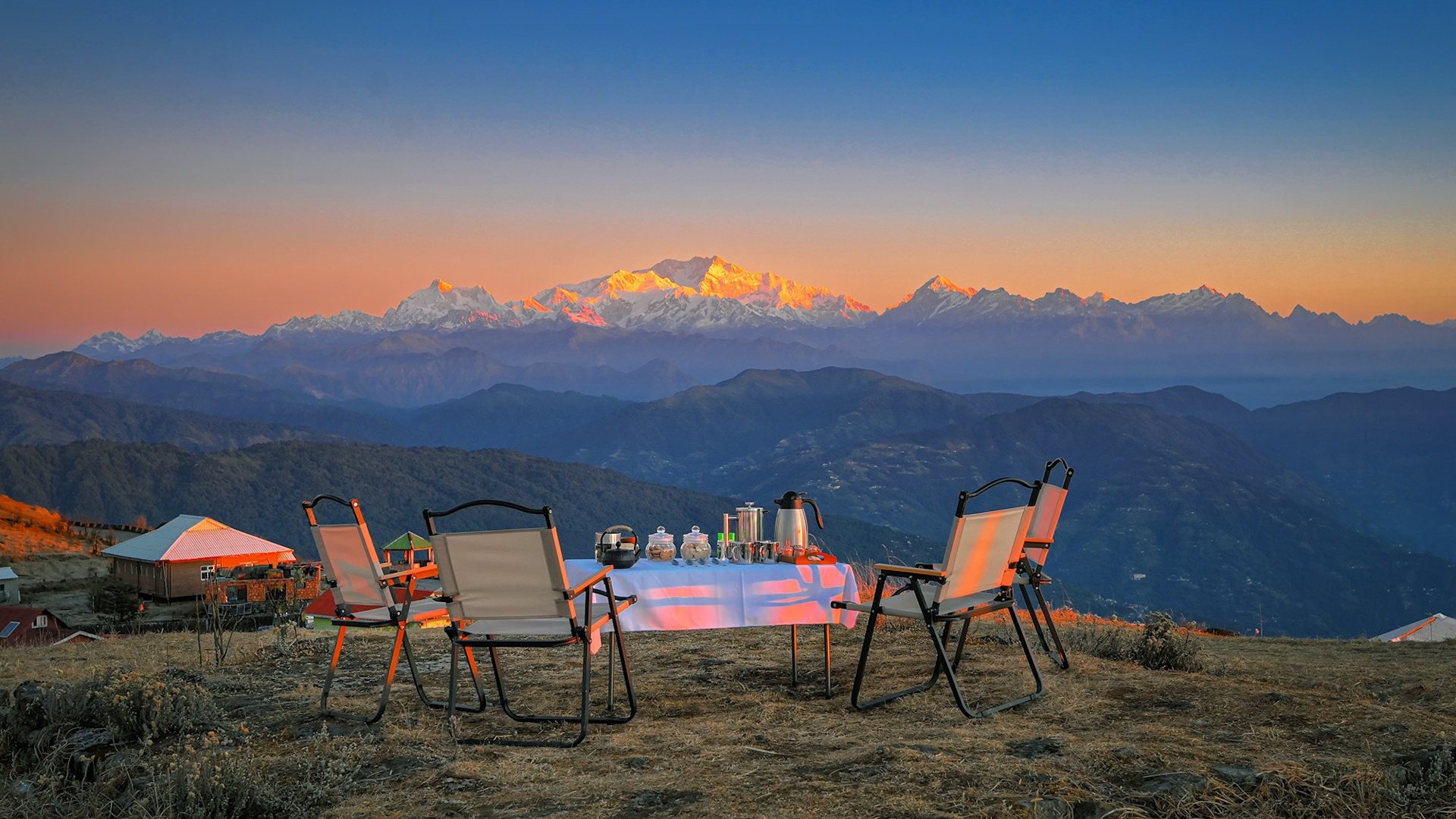 Chairs and a table set out on a ridge at dusk, facing snowy Himalayan peaks