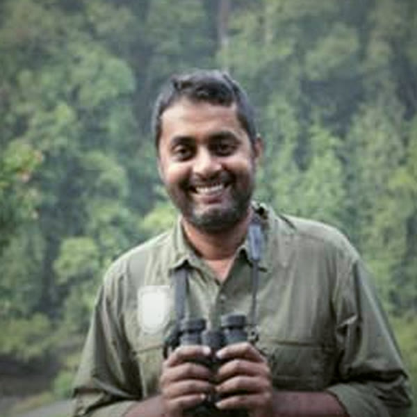 Harsha holding binoculars at the edge of a forest